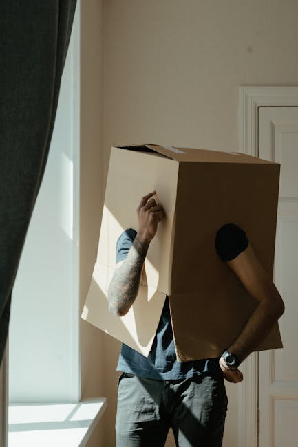 A person with tattoos wearing a casual grey t-shirt and black wristwatch is seen inside a room, carrying a large cardboard box over their head during a home relocation process. The individual's left arm, which is visible, is holding the box, while their right arm is resting on their hip. The room has light-colored walls, with sunlight streaming through a window that is partially covered by a dark curtain, casting shadows on the wall. In the background, a white door with decorative molding is visible. The scene depicts a moving or packing activity, consistent with furniture transport or packing and moving services offered by Hackney Removals, emphasizing the careful handling of household items during house removals.
