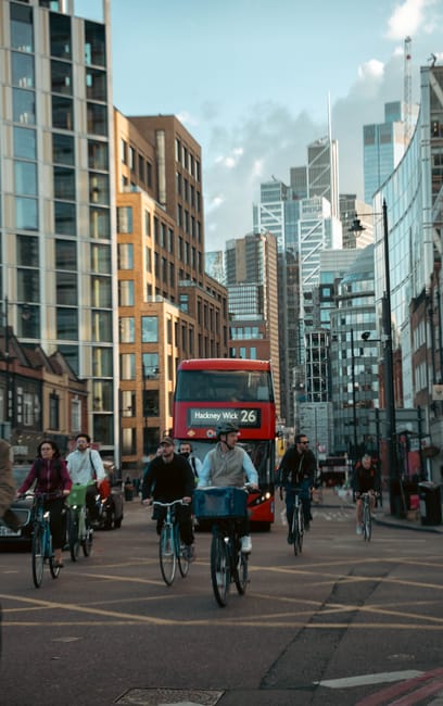 This image depicts a city street scene in Hackney with a group of pedestrians and cyclists navigating the area, including several individuals riding bicycles and walking. In the background, a double-decker bus displaying the route number 26 and the destination Hackney Wick is approaching or stopped near tall modern office buildings and older brick structures, creating a contrast between contemporary and historical architecture. The street is busy with vehicle and pedestrian activity, under a partly cloudy sky with natural daylight illuminating the scene. Although the image focuses on urban transportation and street life, it exemplifies typical environments where home relocation or moving services might be required, with the hustle of city life highlighting the need for efficient packing, furniture transport, and loading processes. Hackney Removals operates within this busy urban context, providing professional moving and removals services that support smooth house relocations amidst such city environments.
