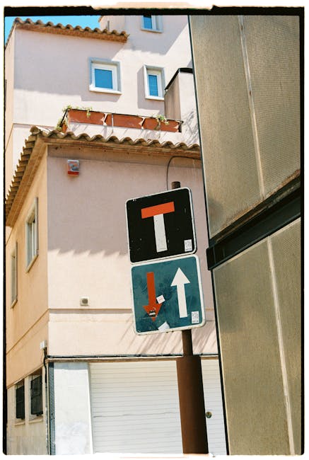 A height restriction traffic sign on a brown metal pole positioned on a narrow street in Dalston E8, showing a black square with a red horizontal bar indicating no through passage for vehicles over a certain height, and a blue sign below with a white upward arrow and a red downward arrow to guide vehicles, with residential buildings in the background featuring white plaster walls, small windows with blue frames, and a tiled roof with overhanging eaves. The scene likely depicts a street where home relocation or furniture transport activities are coordinated, as part of a house removal process, with the signage supporting safe vehicle navigation through narrow urban streets. The lighting suggests a clear day with sunlight casting shadows on the buildings.