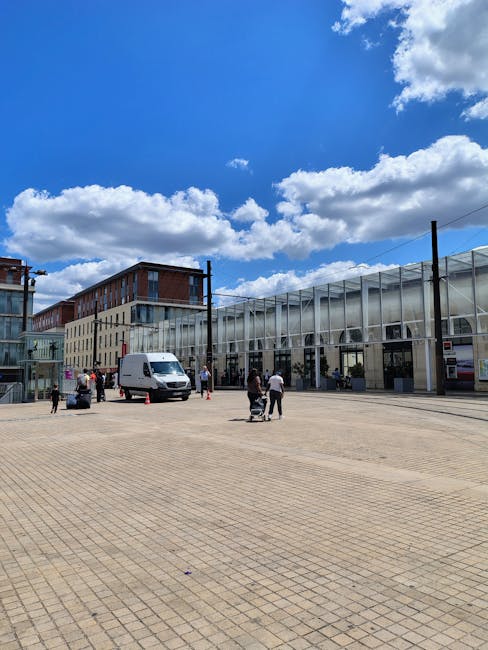 A spacious outdoor area with a paved surface featuring rectangular tiles, adjacent to a modern building with large glass windows and a metallic framework. Several people are present, some standing and others walking, with a few individuals near a white moving van that is parked close to the building entrance. The scene includes cardboard boxes, wrapped furniture, and equipment such as trolleys and straps, indicating a home relocation or furniture transport process managed by Hackney Removals. The sky overhead is mostly clear with scattered clouds, and the environment suggests a daytime setting in an urban location near Olympic Park, aligned with house removal and packing services.