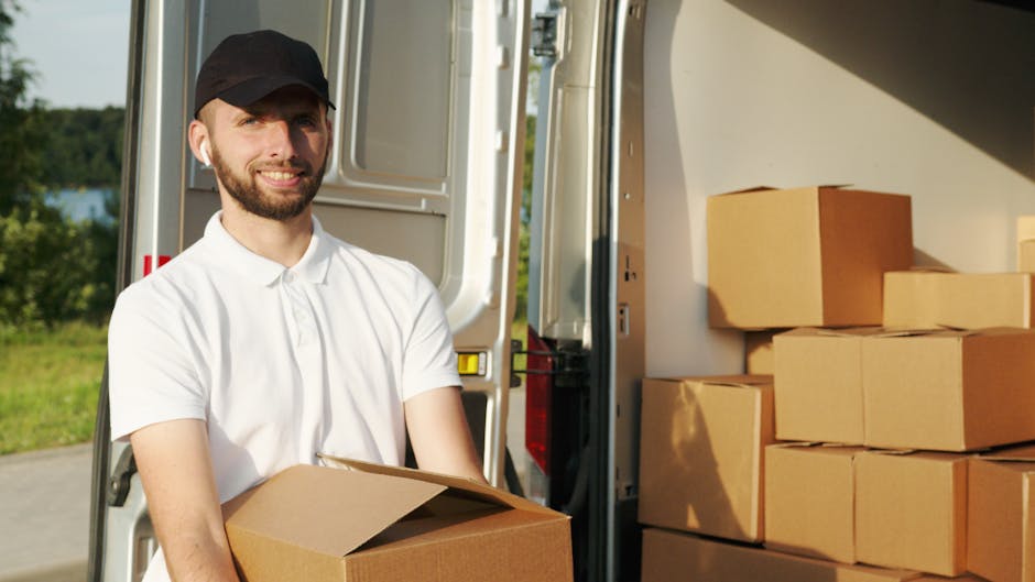 A man with a beard wearing a white polo shirt and a black cap is standing outside during daylight, holding a cardboard box near the back of a moving van. The van's open rear doors reveal a loading area filled with stacked cardboard boxes, some wrapped in plastic, ready for transportation. The moving process appears to be in progress, with the man preparing to load or unload items as part of a home relocation or furniture transport. The background shows a grassy area with trees and a clear sky, indicating an outdoor setting. Hackney Removals’ company vehicles and packing materials are part of the scene, supporting the logistics of packing and moving services. The image captures the practical aspects of a professional removal service, focusing on boxes, packaging materials, and the loading process for a swift and efficient move.