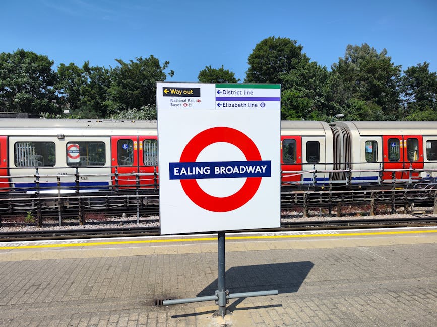 A railway station platform in London with a prominent Ealing Broadway sign mounted on a metal stand in the foreground. Behind the sign, there is a modern train with a silver body, red and blue accents, and multiple windows along its side. The train is positioned on railway tracks with black railings running parallel to the platform edge. To the left, there is an overhead sign indicating directions for the way out, national rail services, and bus connections. The background features lush green trees under a clear blue sky, suggesting a bright, sunny day. The image captures a calm scene related to urban transportation, essential for home relocation or furniture transport services, as provided by Hackney Removals, supporting effective moving logistics and packing and moving processes near Broadway Market.
