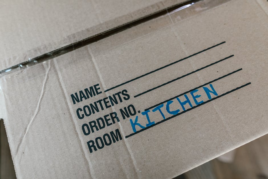 Close-up image of a cardboard moving box with black printed labels indicating sections for name, contents, and order number. The box has a handwritten label in blue ink that reads 'KITCHEN' to specify the contents and designated room, suggesting preparation for a home relocation or packing process. The box appears to be inside a property near the doorway or entrance, possibly during the loading process for a house move. The scene reflects professional packing and furniture transport activities, with materials like cardboard and marked labels used to organise the move. This setup is characteristic of house removals and professional moving services such as Hackney Removals, supporting efficient packing and logistics management in the context of house removals or moving tips near Broadway Market.